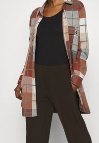 Rutigt flanellskjorta i röd, beige och grå ton; har knäppning fram, två bröstfickor och en avslappnad passform över en svart topp.