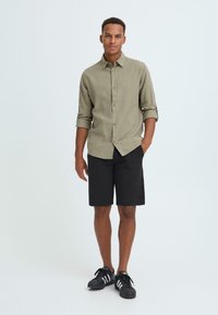 Chemise légère boutonnée vert olive avec manches retroussées, associée à un short gris foncé et des baskets noires à rayures blanches.