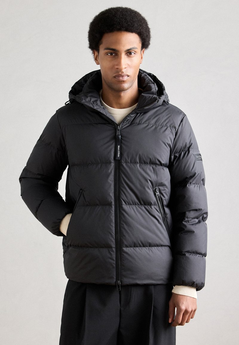 Doudoune noire en matière matelassée élégante avec fermeture éclair, capuche ajustable et poches latérales. Bord-côtes texturé en tricot.