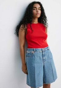 Femme aux longs cheveux bouclés portant un haut rouge sans manches, un short en jean bleu et une ceinture chaîne argentée, debout contre un mur blanc.