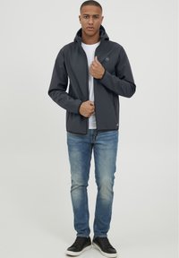 Veste zippée grise avec capuche, arborant un logo sur la poitrine. Associe à un jean bleu et des baskets noires. Matière lisse et légère.