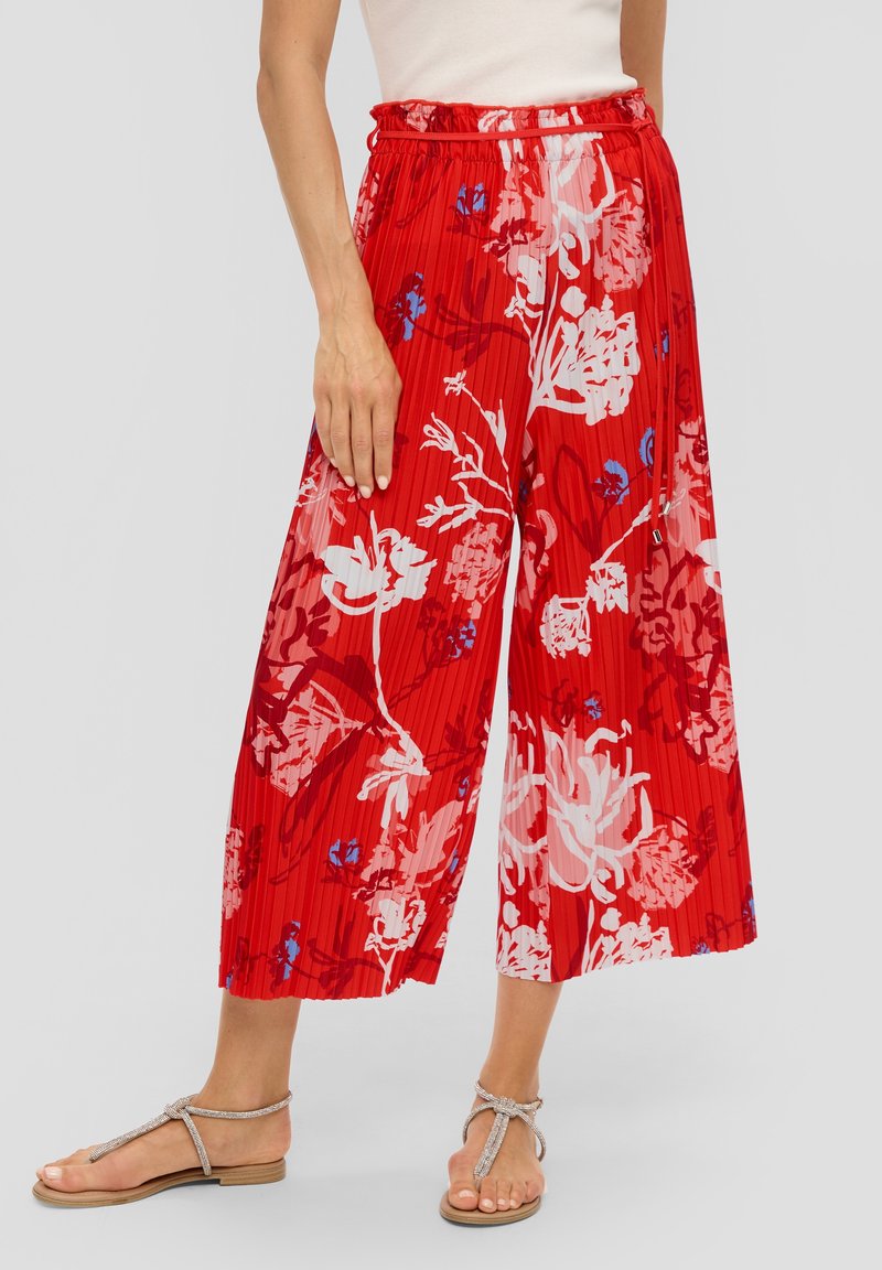 Pantalons larges plissés rouges avec des motifs floraux en blanc et bleu. Taille élastique avec une coupe décontractée. Portés avec des sandales à lanières argentées.