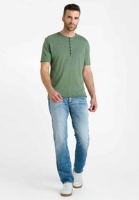 Green short-sleeve t-shirt with a buttoned placket, paired with light blue denim jeans and white sneakers. Casual fit, smooth texture.