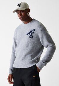Grå strikkegenser med rund hals og marineblå logo på brystet, kombinert med en lys grå caps. Strukturert stoff og avslappet passform.
