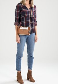 Tan schoudertas met een kettinghengsel, gladde textuur en contrasterend wit paneel. Model draagt een geruite shirt, blauwe jeans en bruine enkellaarsjes.