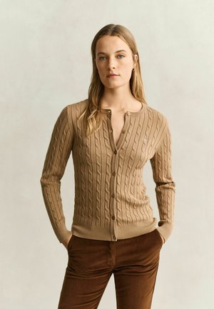 Frau mit langen blonden Haaren trägt eine beige kabelgestrickte, geknöpfte Strickjacke und dunkelbraune Hose, steht mit den Händen in den Taschen vor einem schlichten Hintergrund.