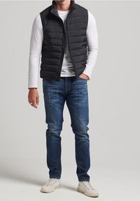 Schwarze gesteppte Weste mit hohem Kragen über einem weißen Langarmshirt, kombiniert mit blauen Jeans und weißen Sneakers. Schlichtes Design, normale Passform.