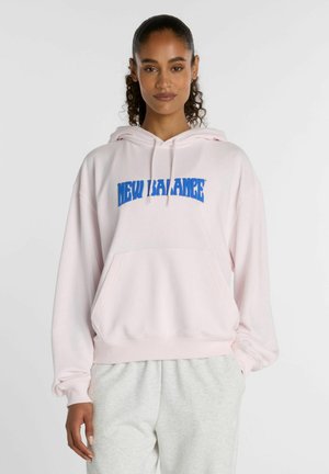 Frau mit lockigem Haar im Pferdeschwanz trägt einen hellrosa New Balance Hoodie und hellgraue Jogginghosen und steht vor einem schlichten weißen Hintergrund.