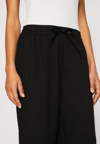 Femme portant un pantalon noir à jambes larges avec taille élastique et cordon, associé à un top blanc sans manches, mains relâchées le long du corps.