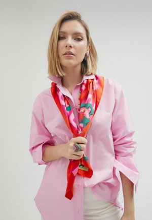 Chemise rose à boutons avec manches retroussées, assortie à une écharpe colorée aux motifs vifs dans des tons rouges et roses, et une bague multicolore ornée de bijoux.