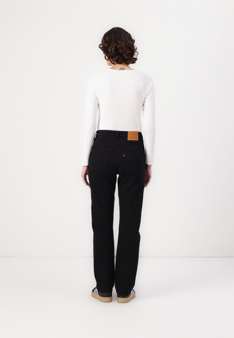 Jeans de mezclilla negra con corte recto, que cuentan con una etiqueta de cuero en la pretina trasera y dos bolsillos en la parte posterior. Acompañados de una camiseta blanca de manga larga.