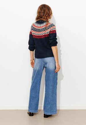Pull en maille bleu marine avec des motifs géométriques multicolores sur le yoke, associé à un jean en denim bleu à jambes larges et des chaussures noires.
