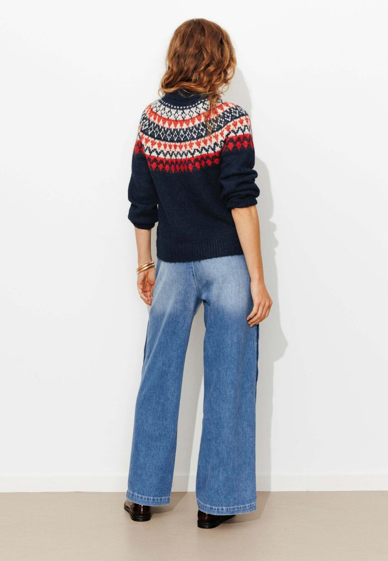 Pull en maille bleu marine avec des motifs géométriques multicolores sur le yoke, associé à un jean en denim bleu à jambes larges et des chaussures noires.