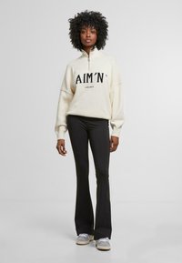 Cream zip-up sweater with black text "AIM’N" and "sportswear" below; paired with black flared pants and gray sneakers.
