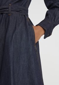 Mörk denimklänning med belagd midja, med sidofickor, kontrasterande orange sömmar och en smidig textur.