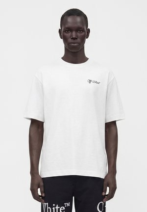 Mand med mørk hud, iført en hvid Off-White t-shirt og sorte bukser med hvide Off-White logoer på en ensfarvet hvid baggrund.