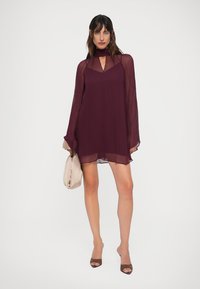 Lühike burgundivari dress code, millel on õhukesed pikad varrukad ja lõikeava kaelus. Kangas näeb välja kerge ja voolav, mida täiendab beež käekott.