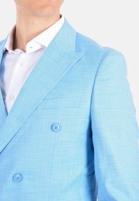 Blazer doppiopetto azzurro chiaro in tessuto testurizzato, con due bottoni rotondi e revers a punta. Monaco della camicia bianca visibile sotto.