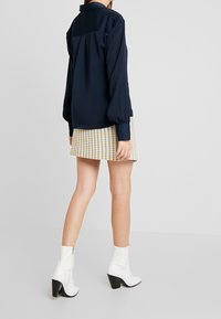 Blusa azul marino de mangas largas, con detalles plisados en la parte posterior, combinada con una mini falda amarilla de cuadros y botas blancas de tobillo con tacones negros.