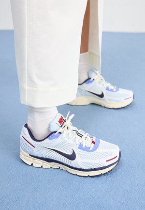 Osoba nosząca białe spodnie, białe skarpetki oraz niebiesko-białe buty do biegania marki Nike stojąca na jasno niebieskim dywanie.