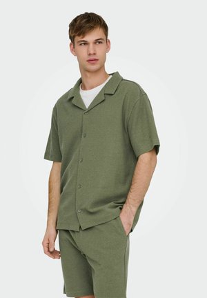 Giovane uomo indossa una camicia a maniche corte verde oliva con bottoni e pantaloncini abbinati con una maglietta bianca sotto, in piedi con una mano in tasca.