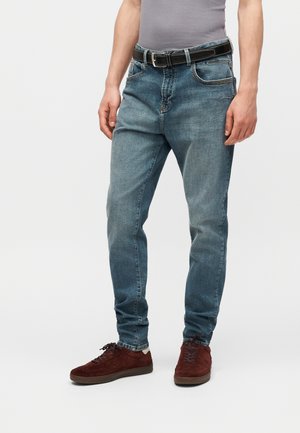 Mies pukeutunut vaaleansinisiin farkkuihin, mustaan vyöhön, harmaaseen toppiin ja tummanpunaisiin mokkanahkakenkiin, seisoo valkoisella taustalla.