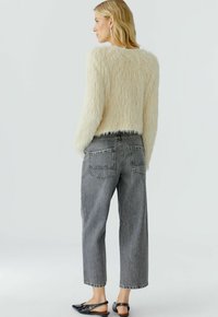 Vrouw in een crèmekleurige, fuzzy trui, hooggewaiste grijze cropped jeans en zwarte slingback flats, met de rug naar de camera op een effen achtergrond.