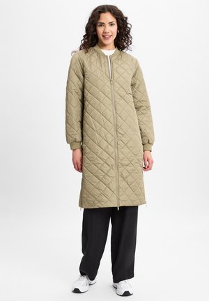 Manteau long olive matelassé avec fermeture éclair à l'avant et poignets côtelés, associé à un pantalon noir décontracté et des baskets blanches, avec un design simple.