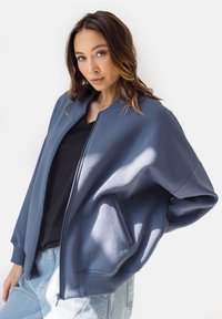 Niebieska kurtka bomberka na zamek, wykonana z miękkiego materiału dresowego; charakteryzuje się luźnym krojem, ściągaczami w mankietach oraz bocznymi kieszeniami. Połączona z czarną bluzką.