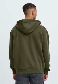 Mannequin in een donkergroene hoodie, met zijn rug naar de camera gekeerd. De achtergrond is licht en eenvoudig, wat de kledingstijl benadrukt.