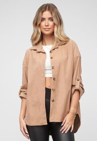 Beige kordfløyelsskjorte i oversized modell med krage, knapper foran og oppbrettede ermer. Kombinert med en hvit topp og svarte leggings.