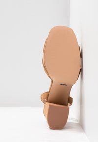 Sandale en daim couleur fauve avec petit talon carré vue de la semelle, appuyée contre l'angle blanc du mur et du sol.