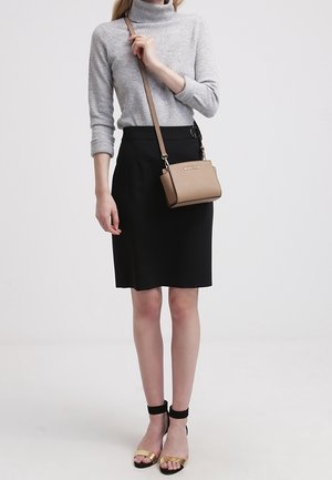 Kožená crossbody taška v tan barvě se zlatými kovovými prvky a strukturovaným designem, umístěná na pozadí šedého svetru a černé sukně.