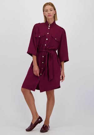 Robe chemise bourgogne à manches courtes, fermeture à boutons, poches à rabat et ceinture à nouer. Texture lisse avec un motif légèrement froissé.