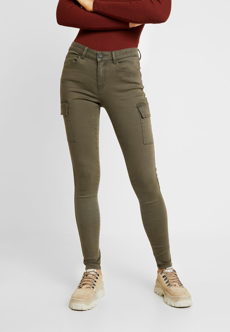 Pantalon cargo slim vert olive avec poches latérales, porté avec des baskets beige épaisses et un haut à manches longues couleur rouille.
