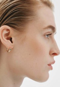 Pendiente de aro dorado con un acento en turquesa, luciendo en un primer plano del perfil de una oreja. Acabado metálico suave, forma circular. Diseño minimalista.
