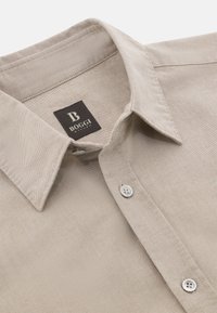 Camisa beige de botones con un cuello estructurado, textura suave y un sutil tejido. La etiqueta dice "BOGGI" en negro.