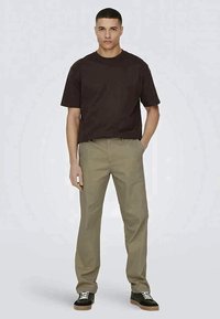 Young man standing with one hand in pocket wearing dark brown t-shirt, beige trousers, and black sneakers with white laces.