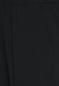 Pantalons noirs en tissu doux, avec une taille large, des plis et des poches latérales. La texture semble lisse et sans texture.