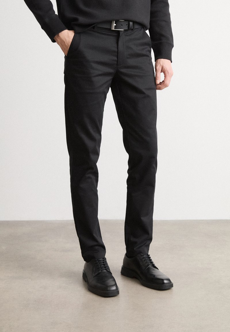 Pantalons noirs à coupe slim, fabriqués en mélange de coton. Présentent des passants de ceinture et des poches latérales, associés à des chaussures noires à lacets.