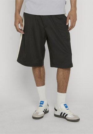 Mand iført sorte knælange shorts med hvide sidelister, hvide sokker og hvide sneakers med sorte striber og blåt logo.