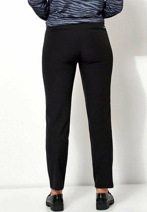 Schwarze, taillierte Hose mit glatter Textur, schmal zulaufenden Beinen und zwei hinteren Taschen. Getragen mit einem gemusterten, dunklen Oberteil und schwarzen Schuhen.