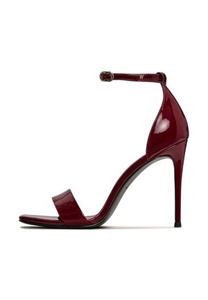 Sandal in vernice rossa con tacco alto, caratterizzata da una singola fascia larga e una fibbia alla caviglia. Presenta una punta appuntita e un tacco sottile.