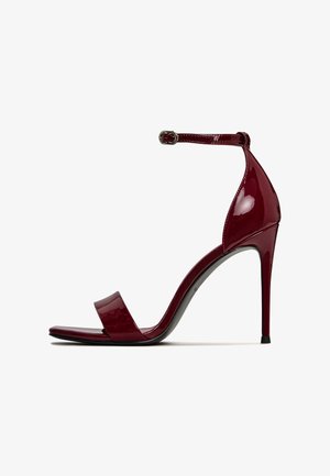 Sandașe cu toc înalt din piele lacuită roșie, cu o curea lată și cataramă la gleznă. Prezentă un vârf ascuțit și un toc subțire de tip stiletto.