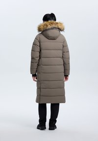 Lång, vadderad kappa i dämpad brun med huva i konstpäls. Har en rak passform och ribbade ärmslut. Bärs med svarta byxor.