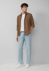 Braune Button-Up-Hemd über einem weißen Grafik-T-Shirt, hellblaue Jeans und weiße Sneaker. Glatter Stoff, lockere Passform und lässiger Stil.