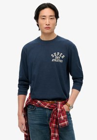 Camisa de manga larga de color navy con el logo "SUPER DRY ATHLETICS", combinada con una camisa de franela a cuadros atada a la cintura y jeans de mezclilla. Accesorio de reloj marrón.