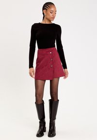 Czerwona zamszowa mini spódnica z rozkloszowanym krojem, ozdobiona trzema metalowymi guzikami, zestawiona z czarną bluzką z długim rękawem i czarnymi botkami do kolan.