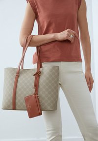 Beige patterned tote bag with brown leather handles, featuring a small matching zip wallet attached. Model wearing a rust-colored top.