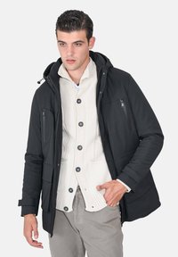 Giacca esterna nera con cappuccio, con una tasca con cerniera, indossata sopra un cardigan crema con grandi bottoni e un motivo lavorato a maglia.